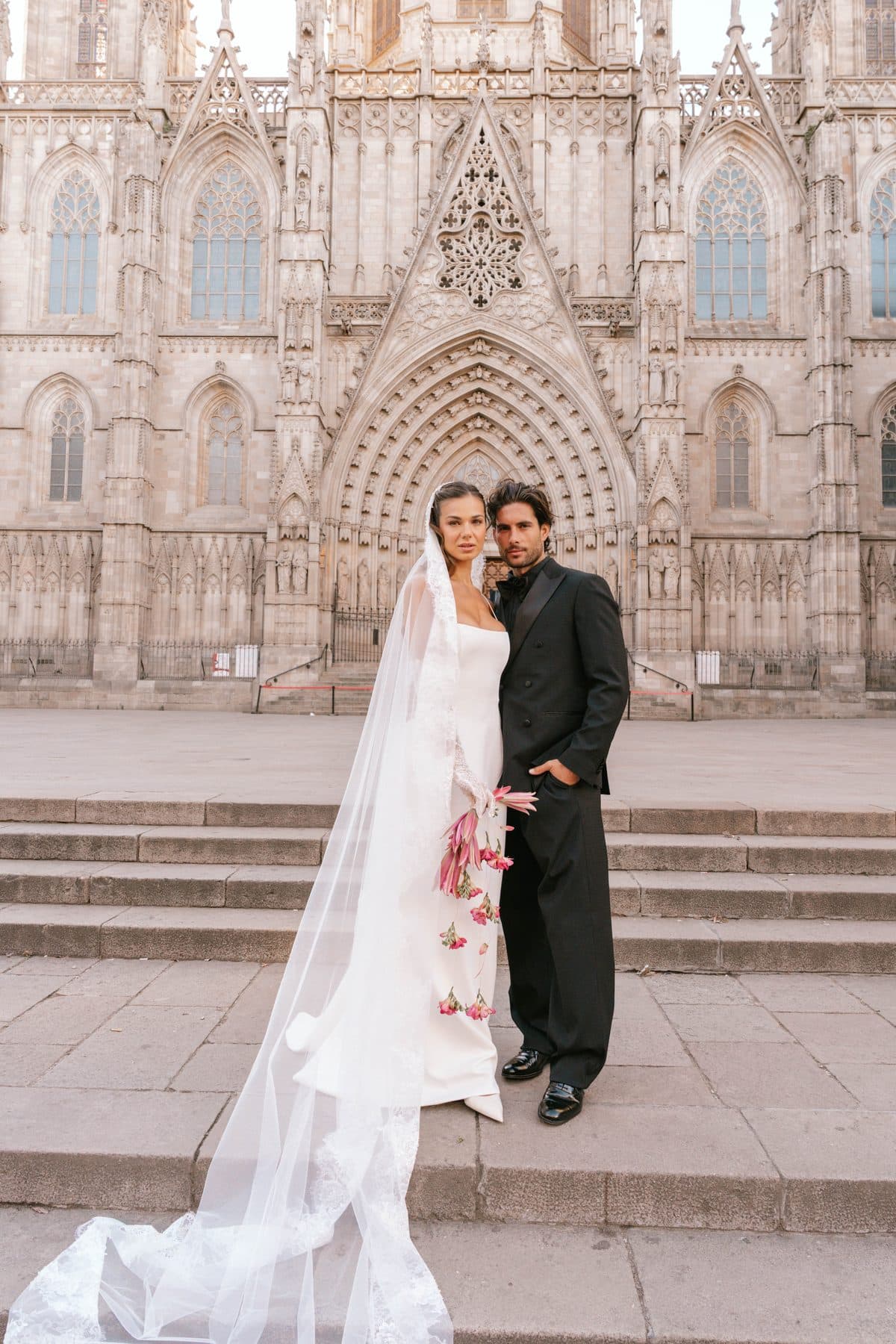 Bride in a lace veil and groom in a black tuxedo on the steps of a Gothic cathedral